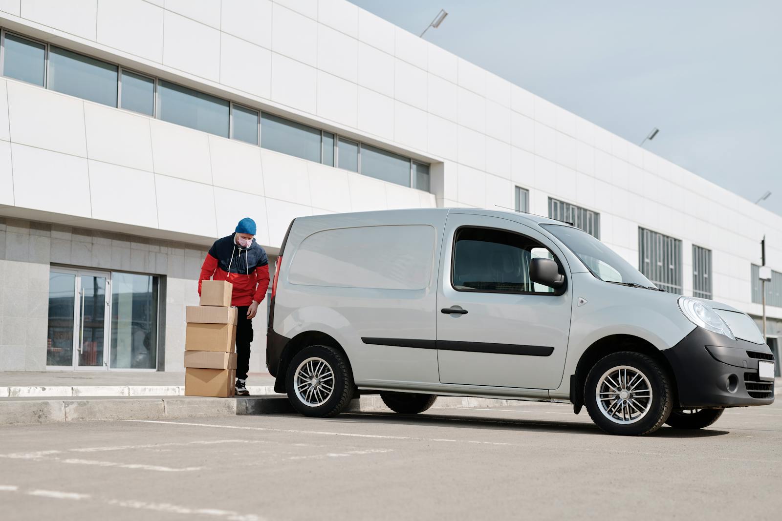 A delivery driver wearing a face mask unloads packages from a van outside a modern building.